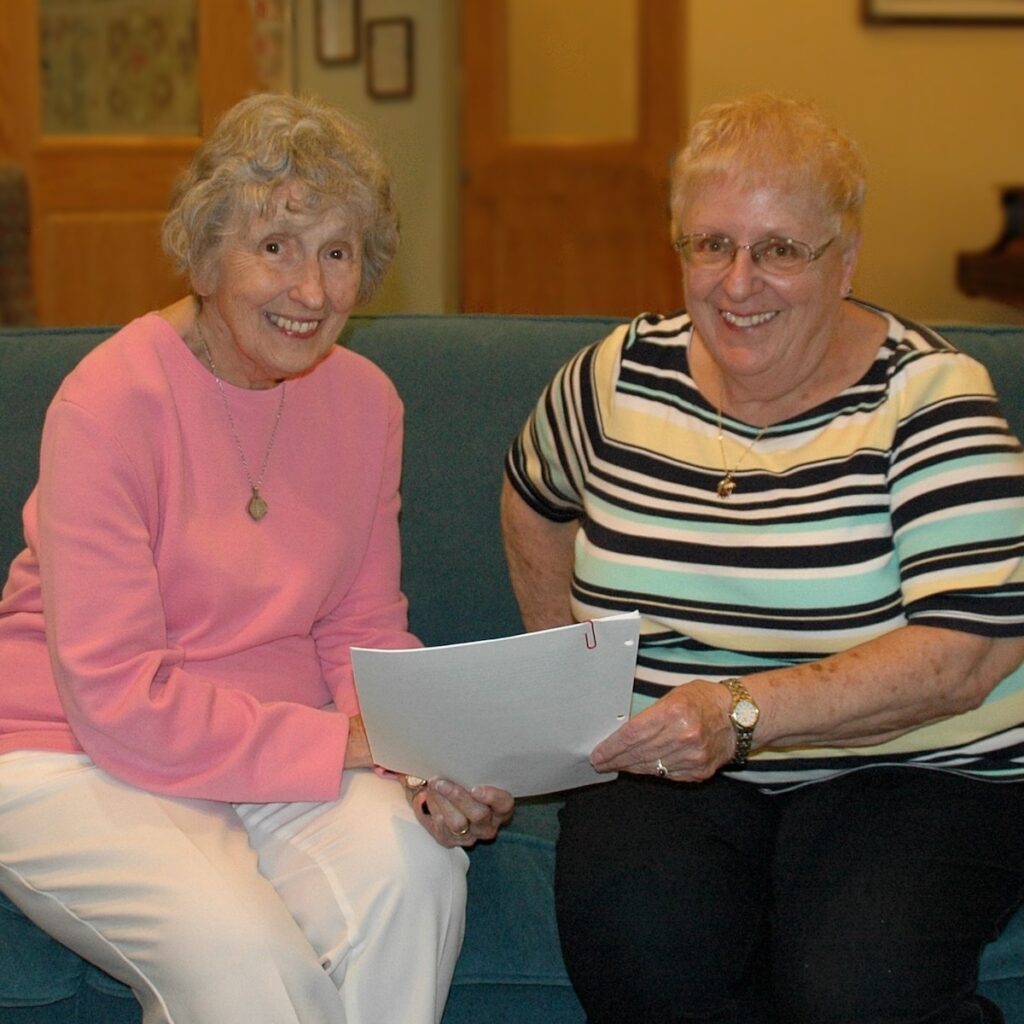 Two women from the Benedictine Sisters sit side by side holding a sheet of paper between them.