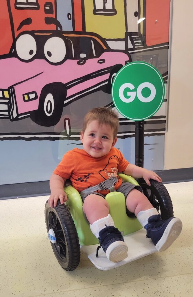 A little boy in an orange shirt sits in a green small wheelchair in front of a cartoon car painting on a wall.