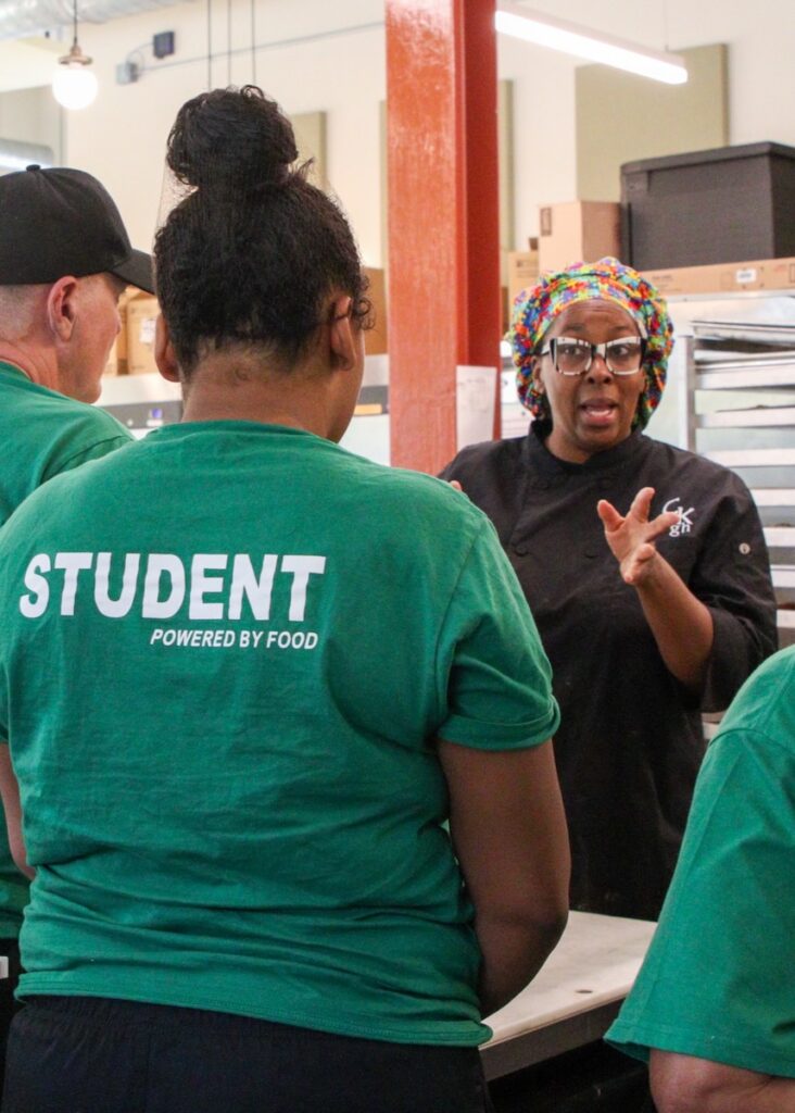 A student in a green shirt watches a professional woman chef.