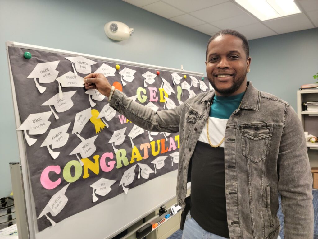 A black man pinning a paper graduation hat onto a cork board with several others.