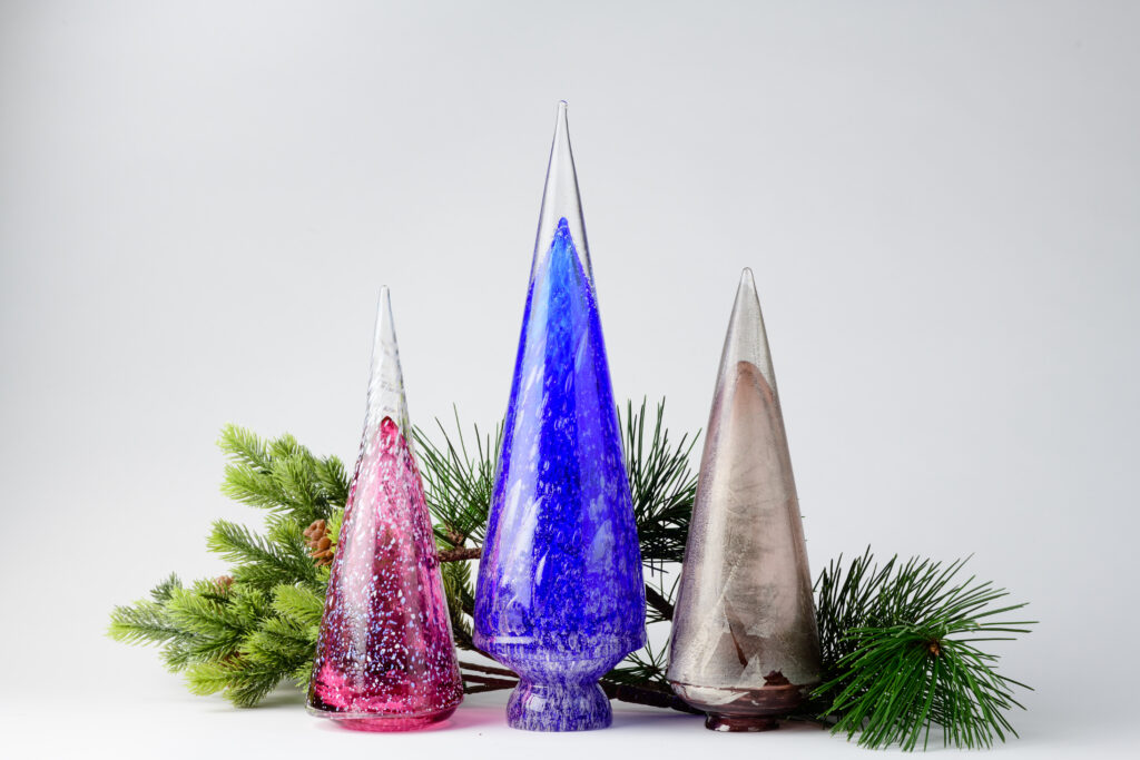Three colorful, glass trees in front of a pine tree branch.