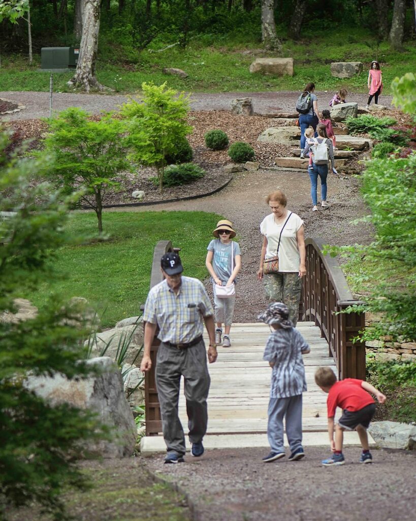 Various people of all ages and kids walk across a bridge and through the scenery at the Pittsburgh Botanic Gardens.