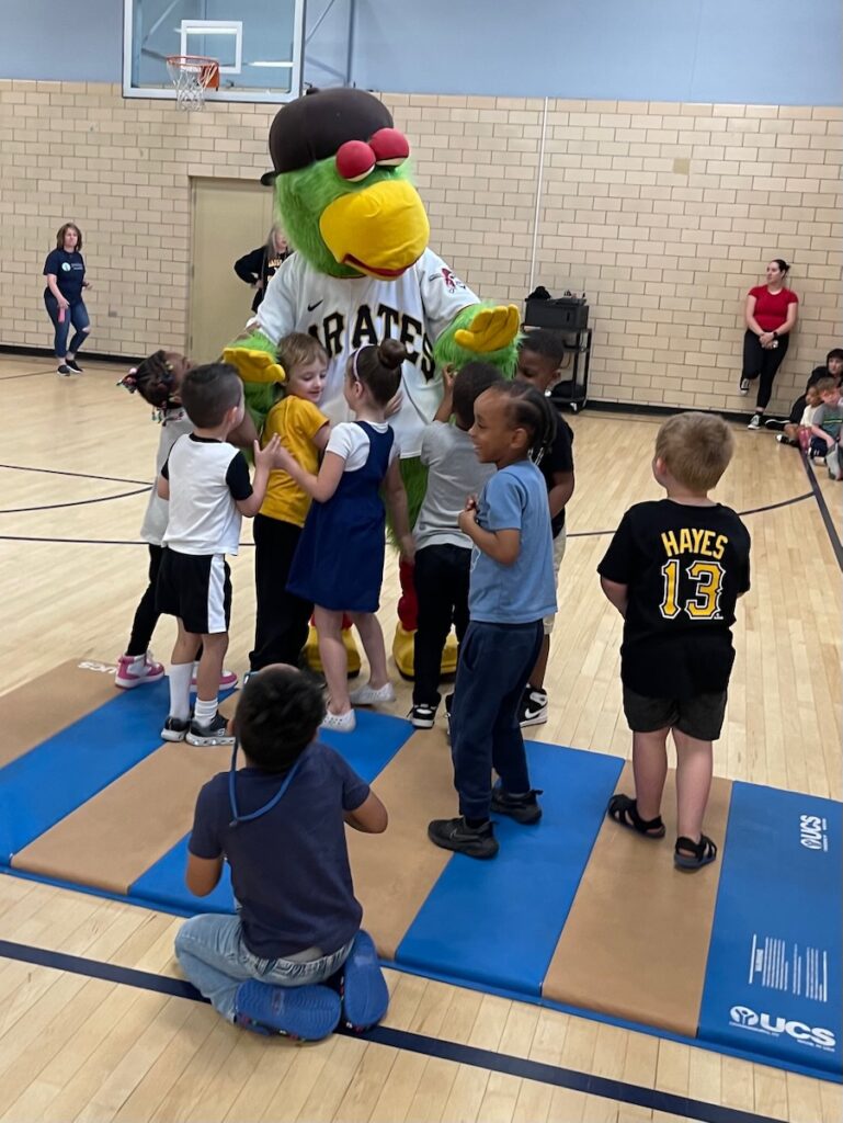 A group of little kids surround the Pirates parrot mascot as some hug him and others stand back in a gym.