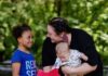 Giving Guide 2024: Trying Together A woman in a black shirt holding a baby leans down to speak to a young girl in a blue shirt who is holding on to a pink pinwheel.