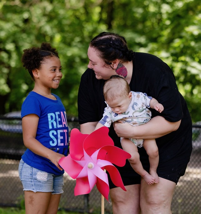 A woman in a black shirt holding a baby leans down to speak to a young girl in a blue shirt who is holding on to a pink pinwheel.