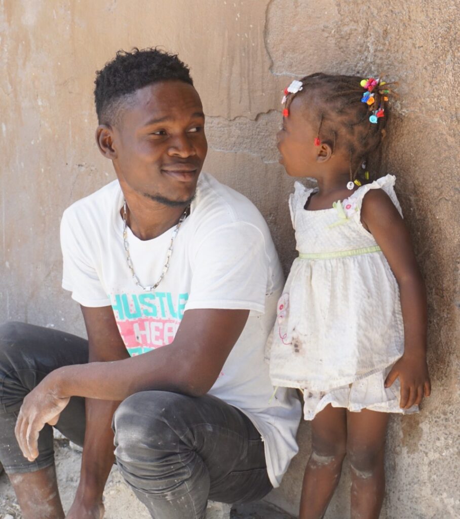 A Hatian man in a white shirt kneels down against a wall next to a young girl in a white blouse and shorts.