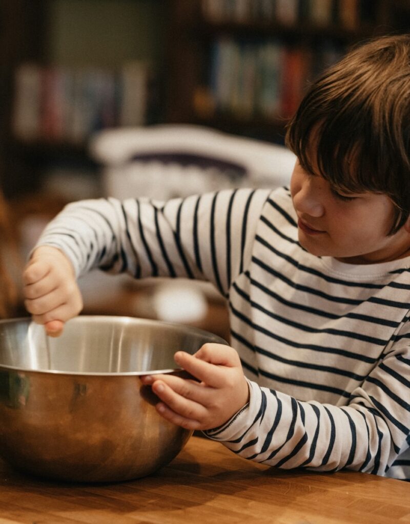 A young boy in a striped sweater cracks an egg into a metal bowl on a wood countertop.