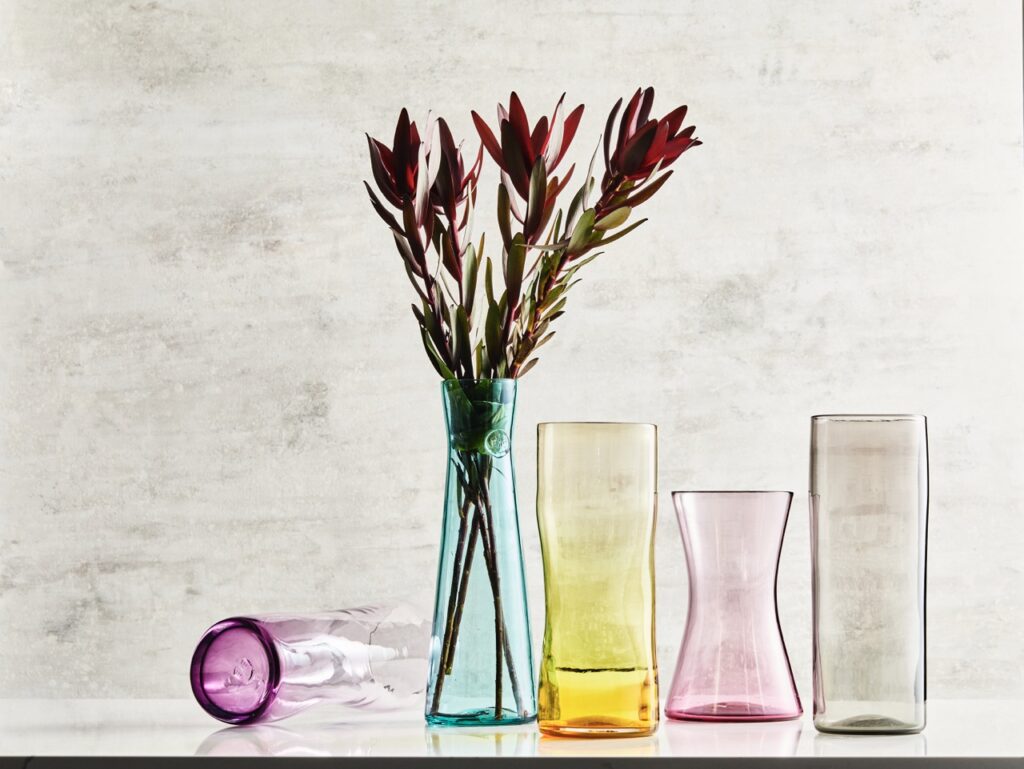 Colorful glass vases for the home in colors purple, blue, yellow, pink, and black sit on a beige background.