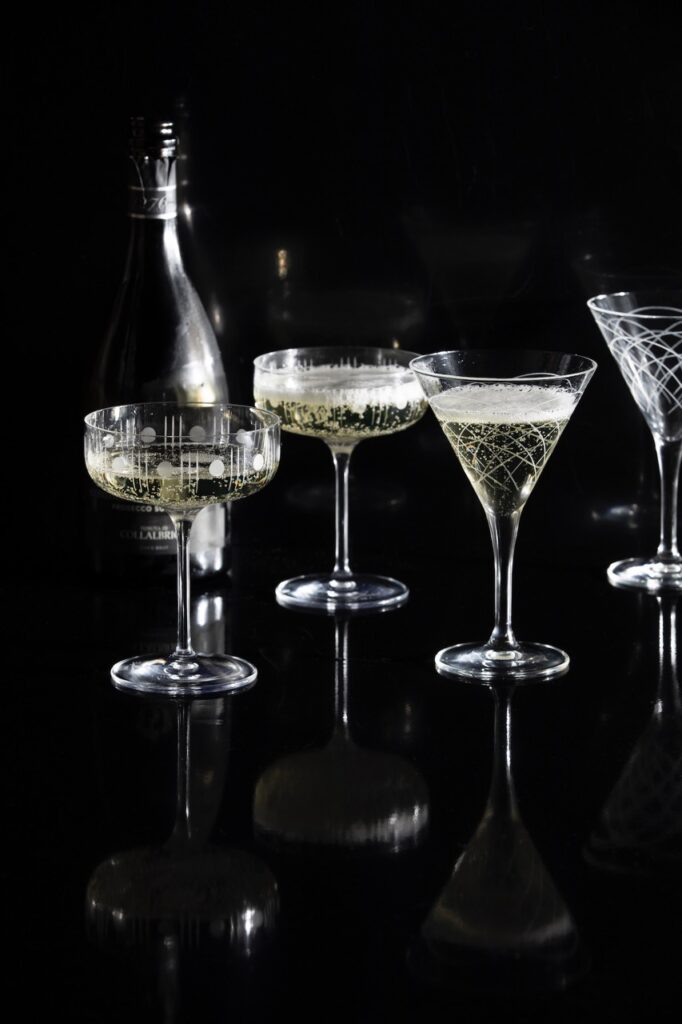 A set of tall glass cocktail glasses in two different styles sit with bubbly liquid in them on a black background.