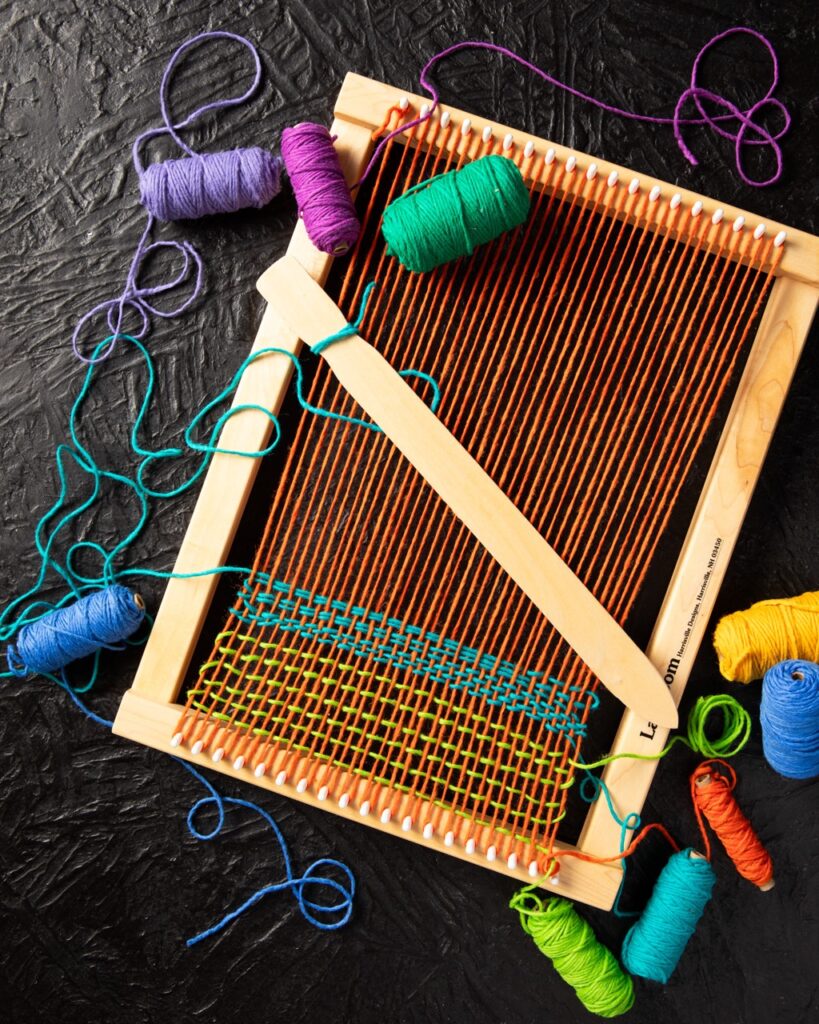 A weaving board sits on a black table in a brown color as purple, yellow, and blue thread is woven into the board.