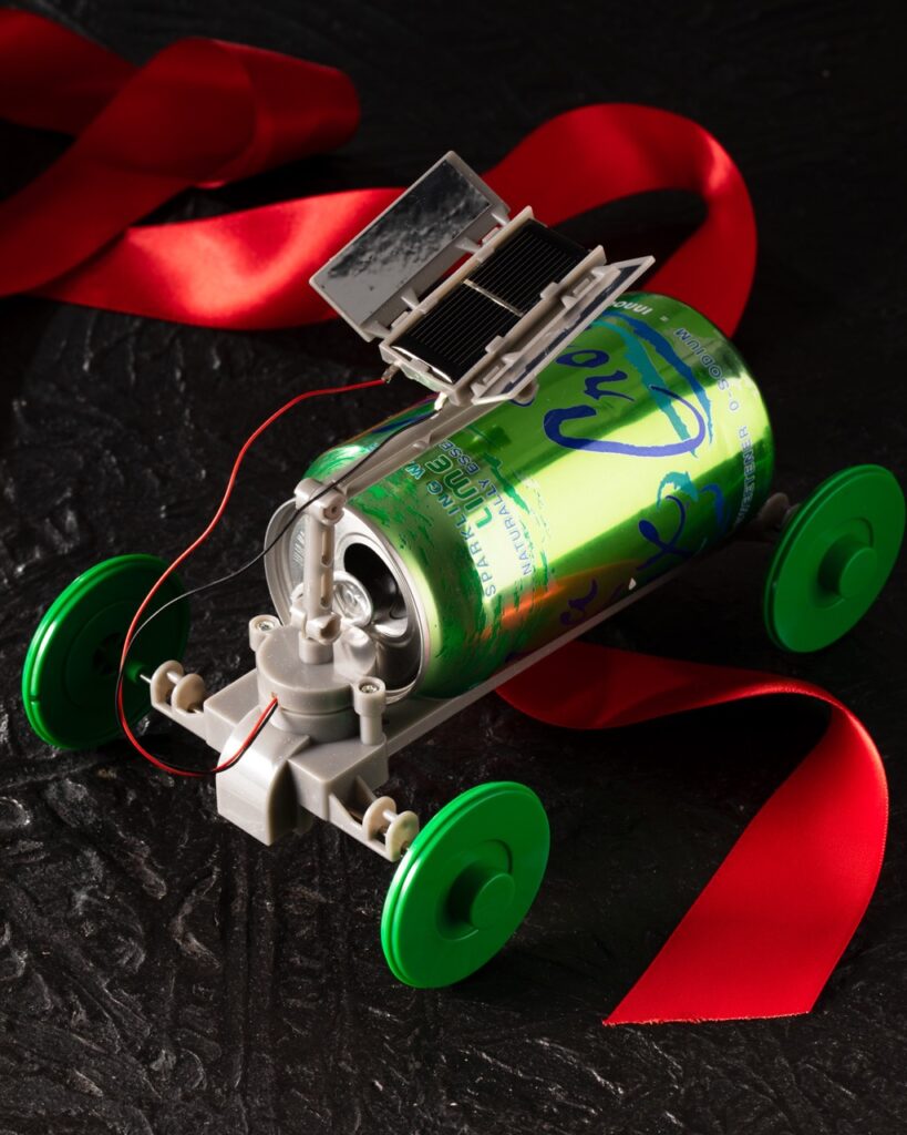 A small electric kids car toy made out of a green soda can and with green tires sits on a table with a red ribbon running under it.