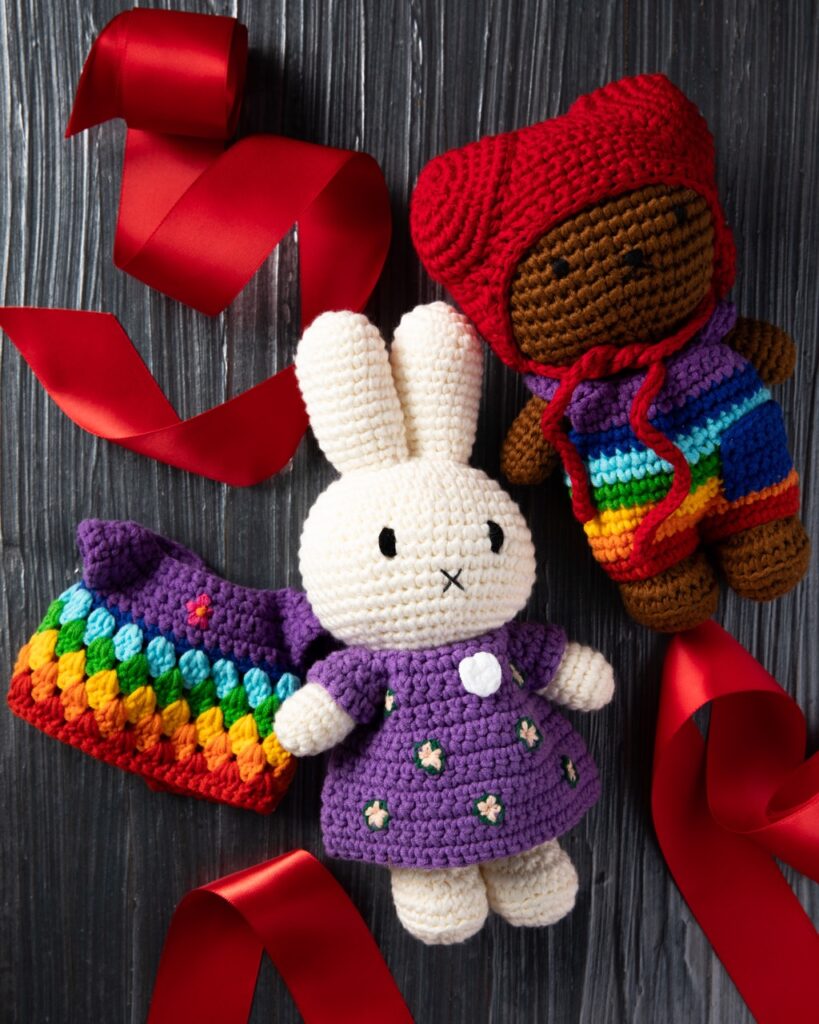 A small crochet bunny with a purple dress sits in the middle beside a brown bear crochet animal in a rainbow jumpsuit and red hat. Red ribbon sits under the kids stuffed animals.