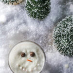 A Melted Snowman cocktail with an edible face sits in the middle of snow and trees.