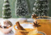 Ginger Snap Cocktail Two clear glass mugs with a ginger snap colored beverage garnished with a cinnamon stick, star anise, and lemon on a snowy surface with a shiny Christmas bulb and little pine trees in the background.