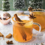 Two clear glass mugs with a ginger snap colored beverage garnished with a cinnamon stick, star anise, and lemon on a snowy surface with a shiny Christmas bulb and little pine trees in the background.