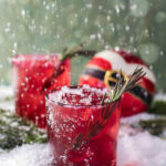 Two red rocks glasses with a spring of rosemary garnish, sitting on a snowy surface with a round Christmas ball that. looks like Santa's outfit.