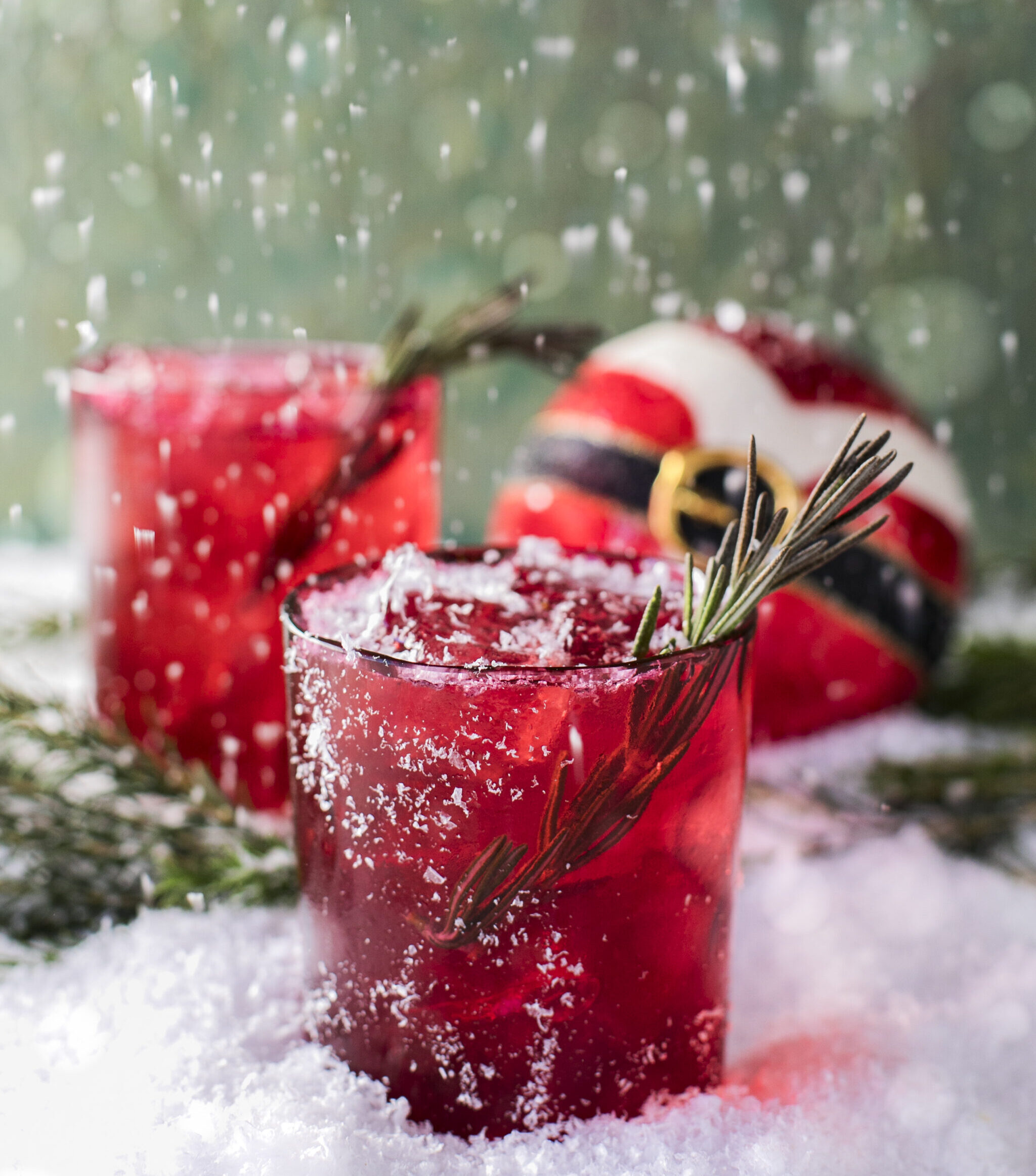 Two red rocks glasses with a spring of rosemary garnish, sitting on a snowy surface with a round Christmas ball that. looks like Santa's outfit.