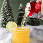 A red can of beer pours into a glass of Cousin Eddie's Trailer Fuel surrounded by snow and trees and garnished with a lime wheel.