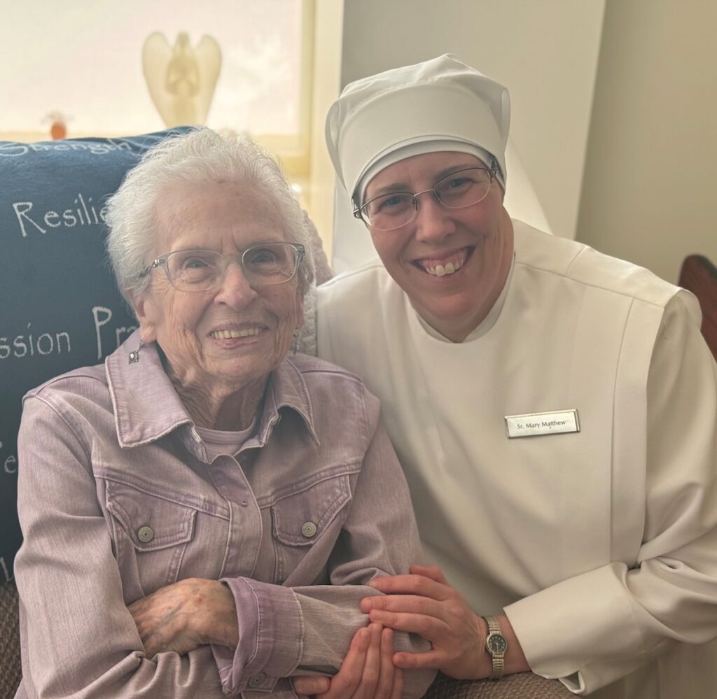 A woman in a whit nurses outfit from the Little Sisters of the Poor holds an elderly woman's arm.