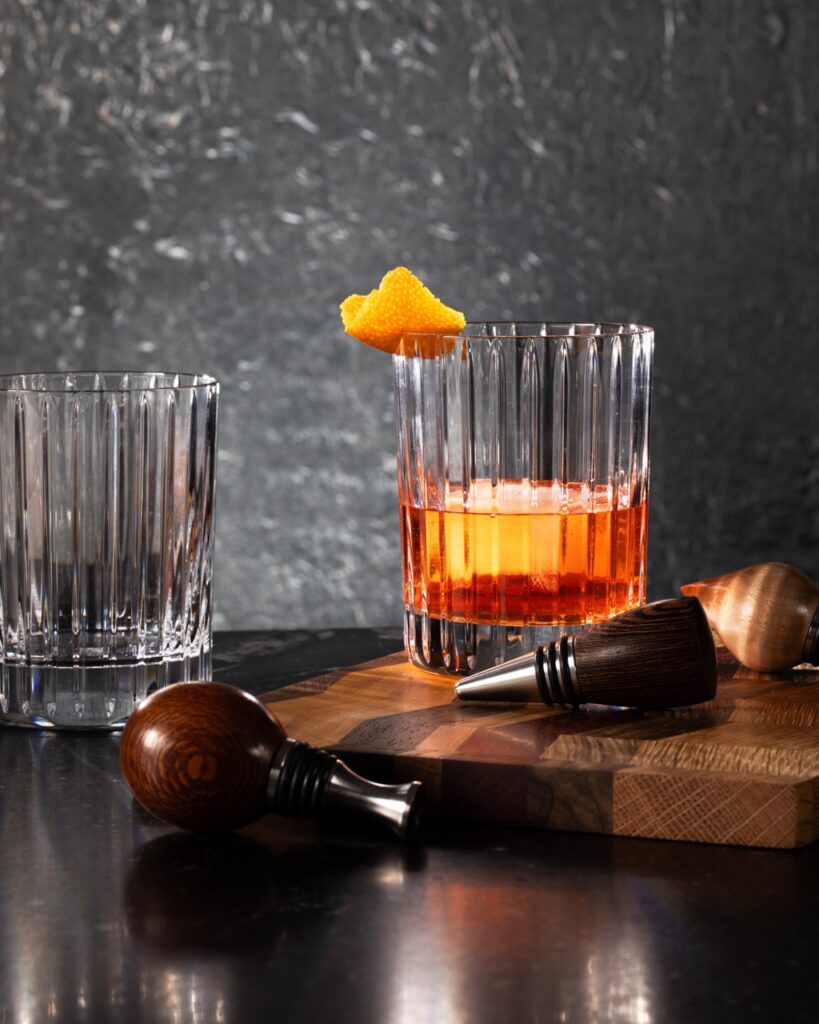 A cocktail glass with an orange drink instead and orange garnish sits behind two wood top wine stoppers as another empty glass sits off to the left.