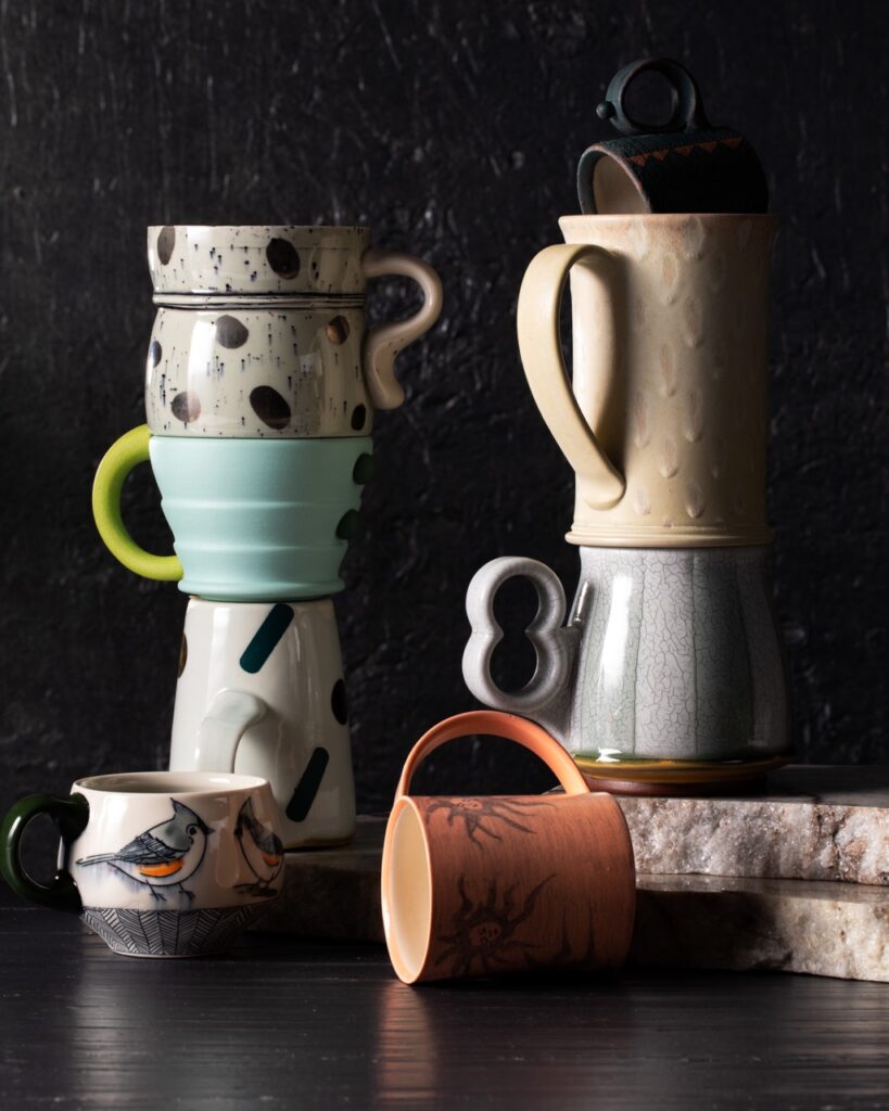 Handcrafted pottery mugs sit in two stacks on a black table while a knocked over brown mug lays in front of the two stacks.