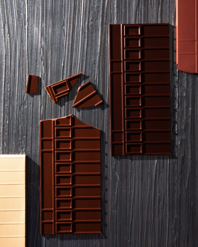 A white chocolate, milk chocolate, dark chocolate, and caramel bars sit diagonally from each other on a grey table. The milk chocolate bar has the top broken into pieces.