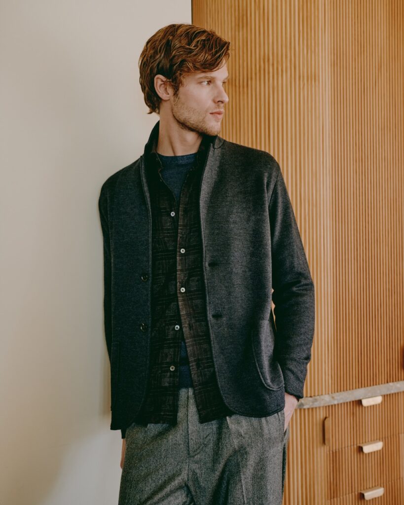 A man leans against a beige wall wearing a mens earthy toned button up shirt, jacket, and green plaid pants.