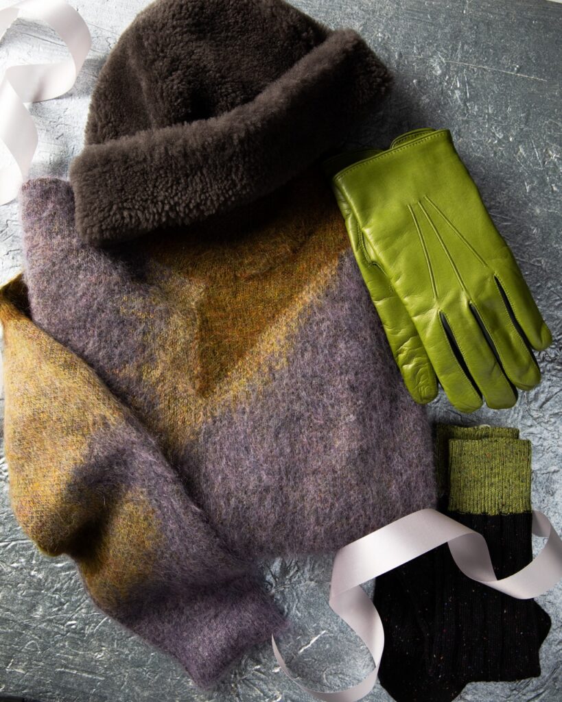 A men's purple and tan sweater sits on a table with a dark brown fuzzy mens hat, green mens gloves, and black and green socks.