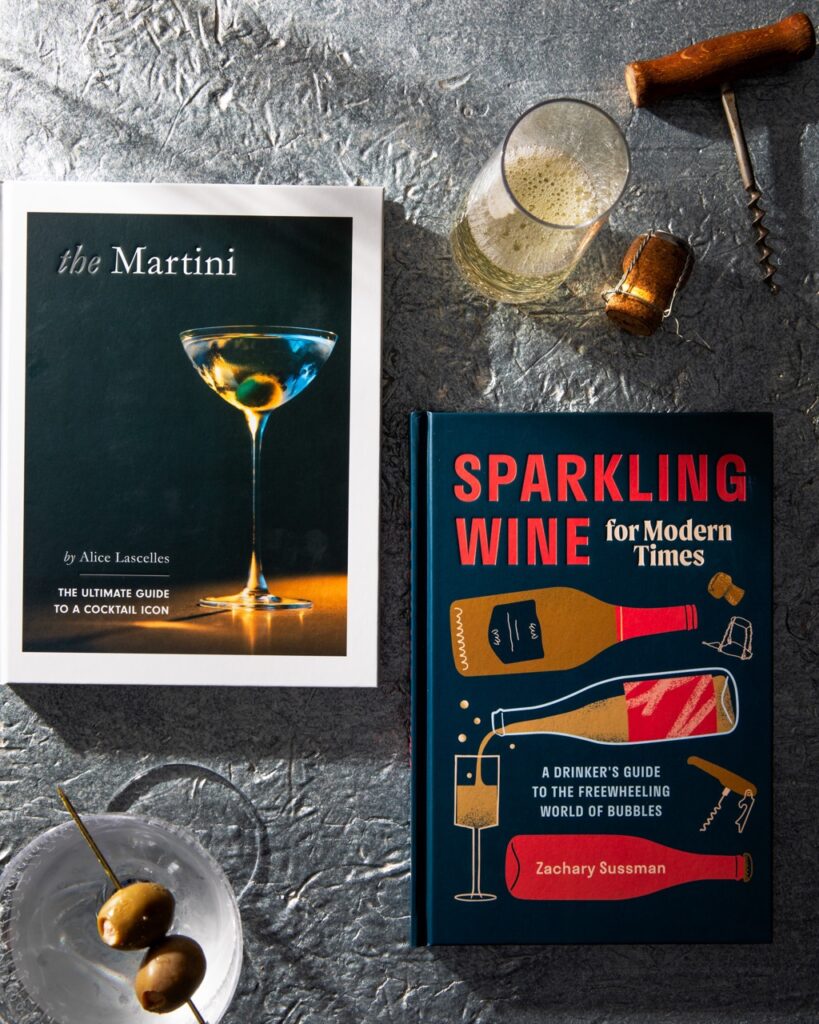 Two wine books, one about martinis, one about sparkling wine, sit on a grey table with a martini sitting beneath one book and a champagne glass above the other.