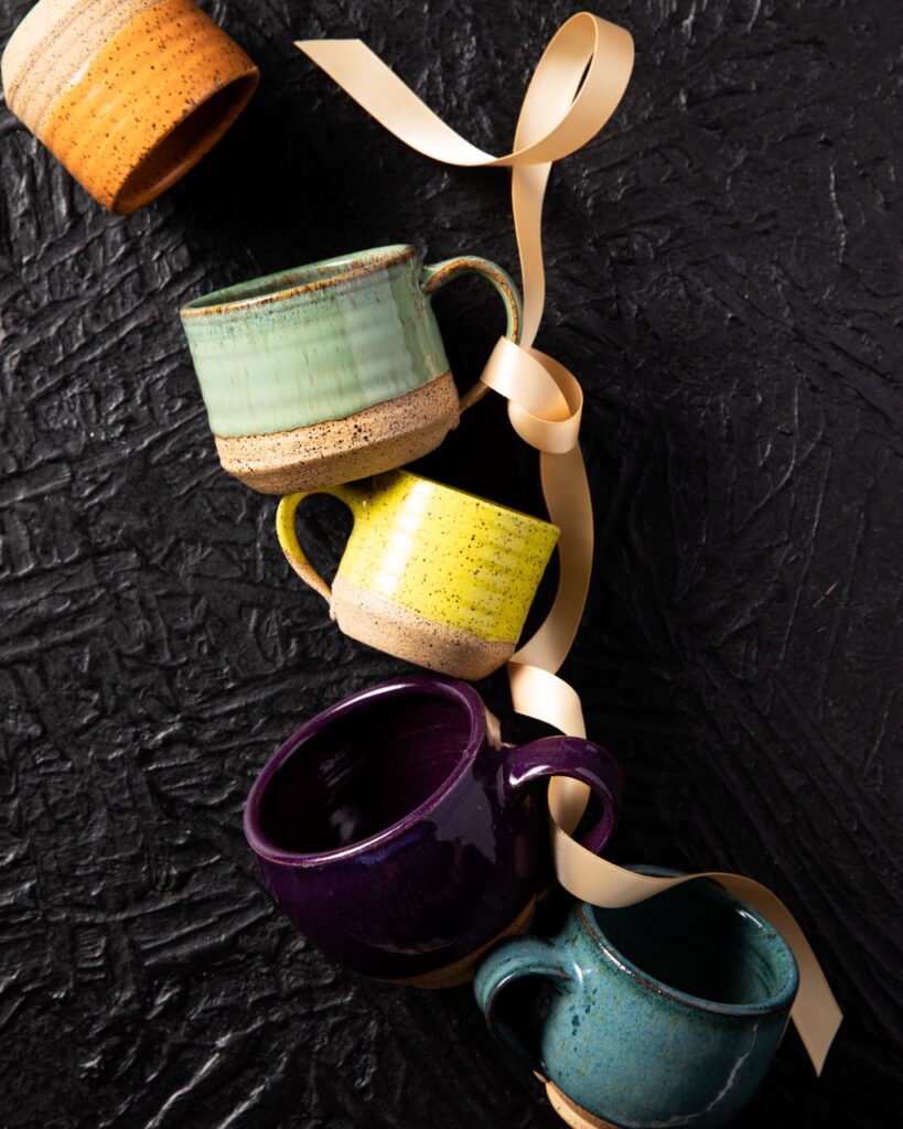 An orange, green, yellow, purple, and blue mugs sit in a line, their handles laced through a pale ribbon.