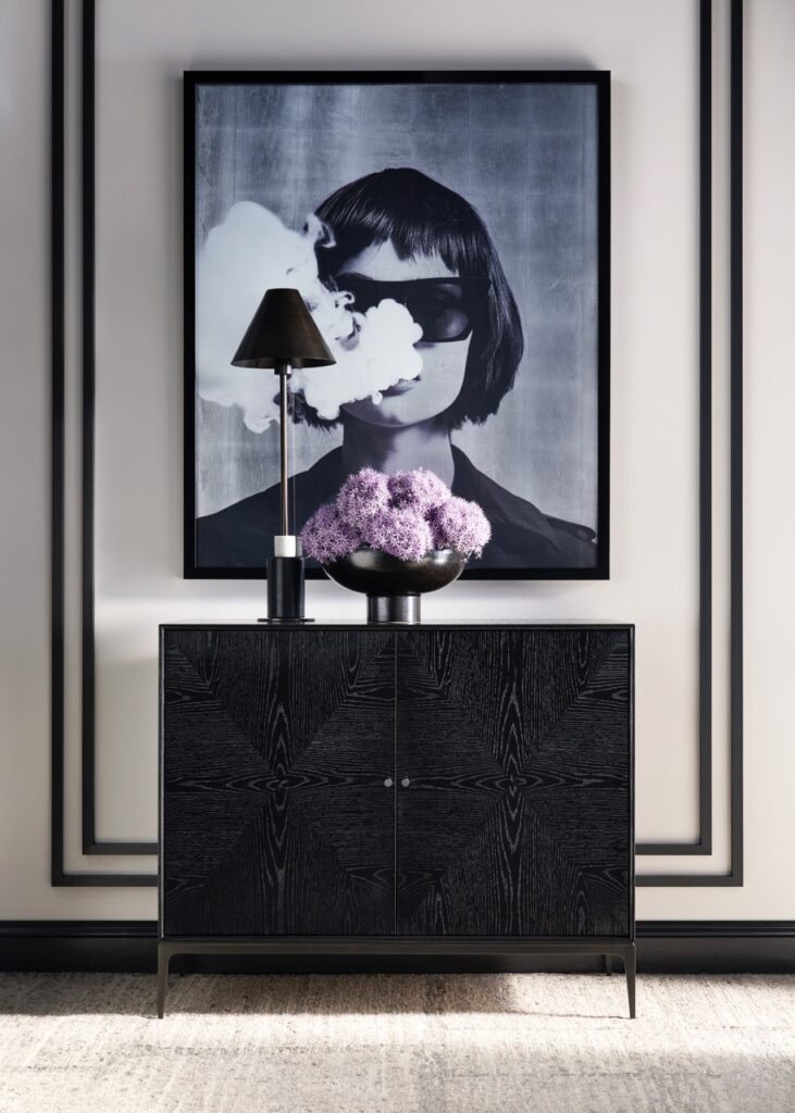 A portrait of a woman with her eyes blacked out sits behind a black cabinet with a lamp and purple flowers on top.