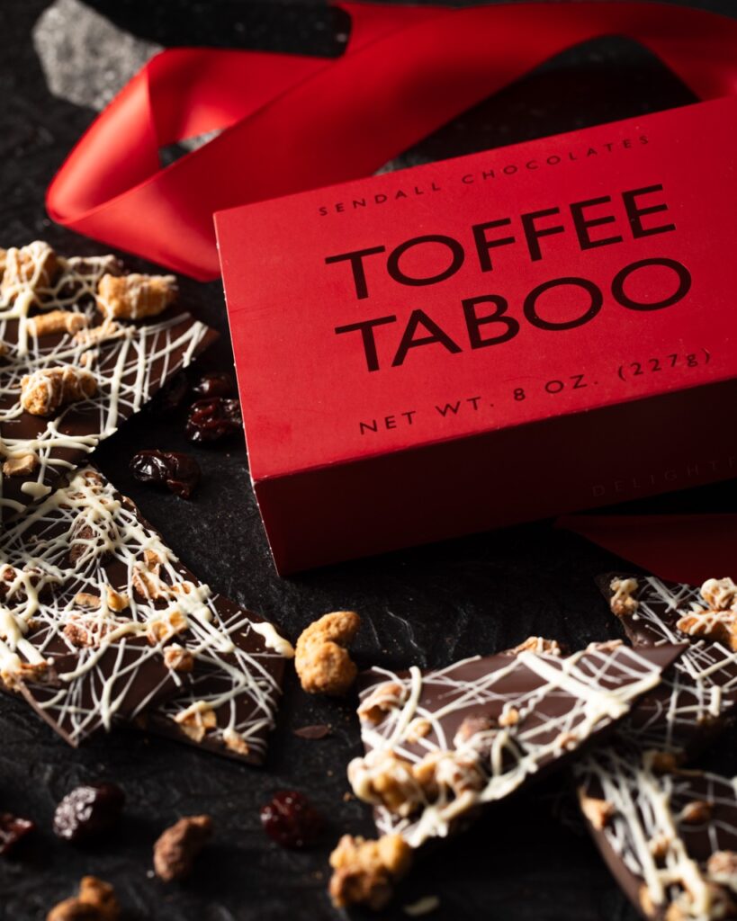 A red box of toffee taboo sits behind various pieces of broken chocolate bark with a white drizzle on top.