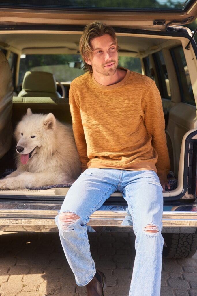 A man sits in the back of a car in an orange men's sweater and jeans while a white dog sits beside him.