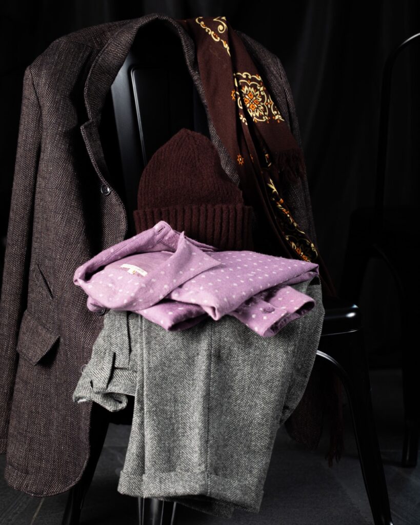 A stack of men's clothing with a pair of grey pants and a purple shirt sits on a chair with a brown suit men's jacket draped over it.