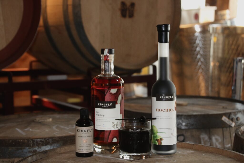Three bottles of Nocino Rye in front of distillery barrels and beside a glass of the drink.