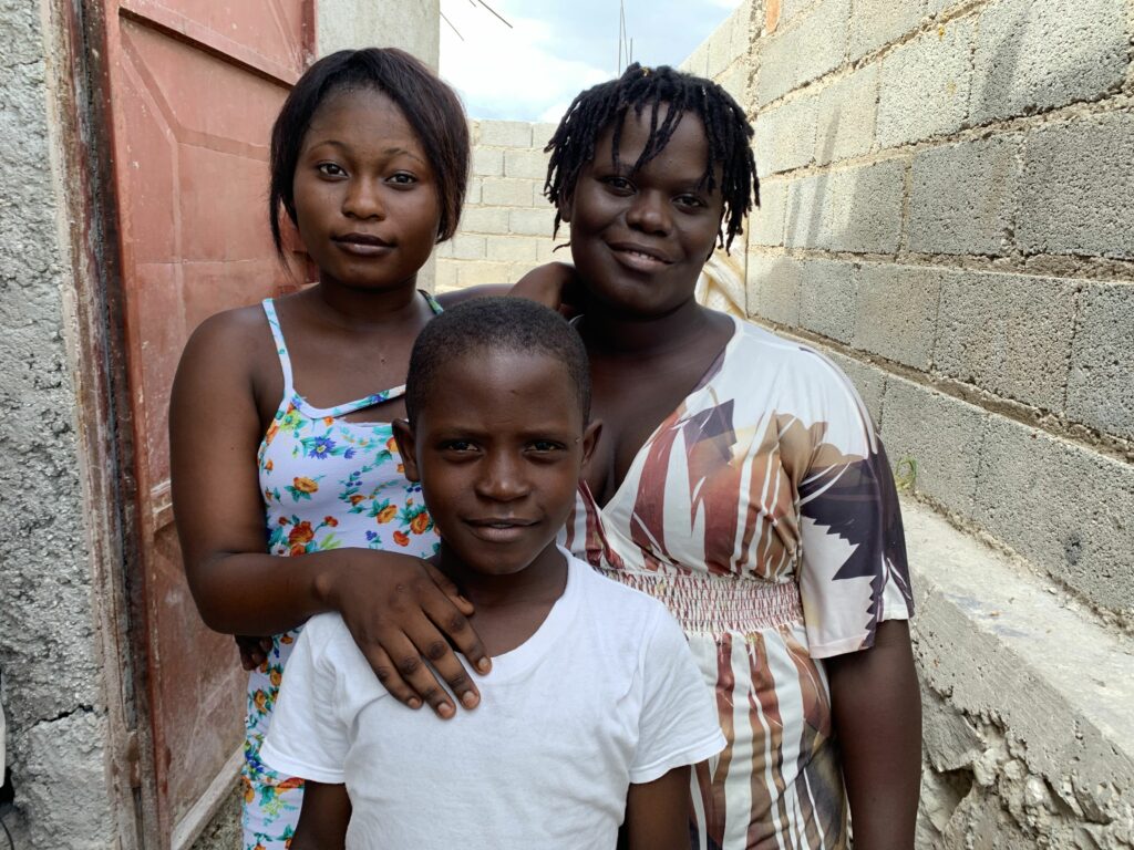 A Haitian woman with her teenage daughter and younger son, standing between block wall structures, smiling for the camera.
