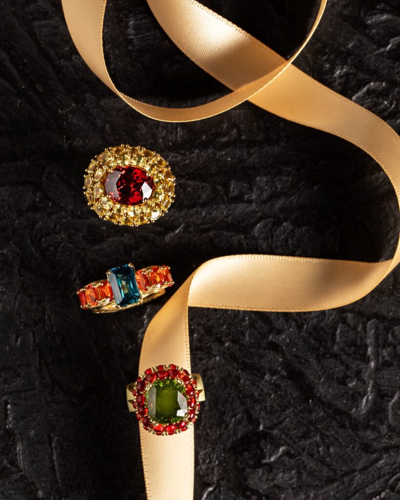 On a string of gold ribbon sits three womens rings in red, orange, blue, and gold gem tones.