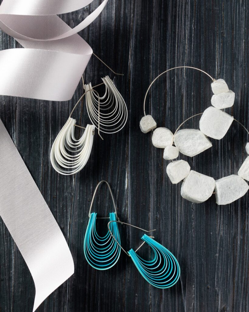 Three sets of earrings made of wire and beads in blue and two in white sit on a black background with a white ribbon cascading down the left side.