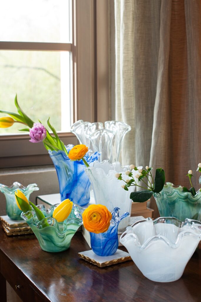 Various glass bowls in green, blue, and clear sits on a brown table, each has a different colored flower in them.