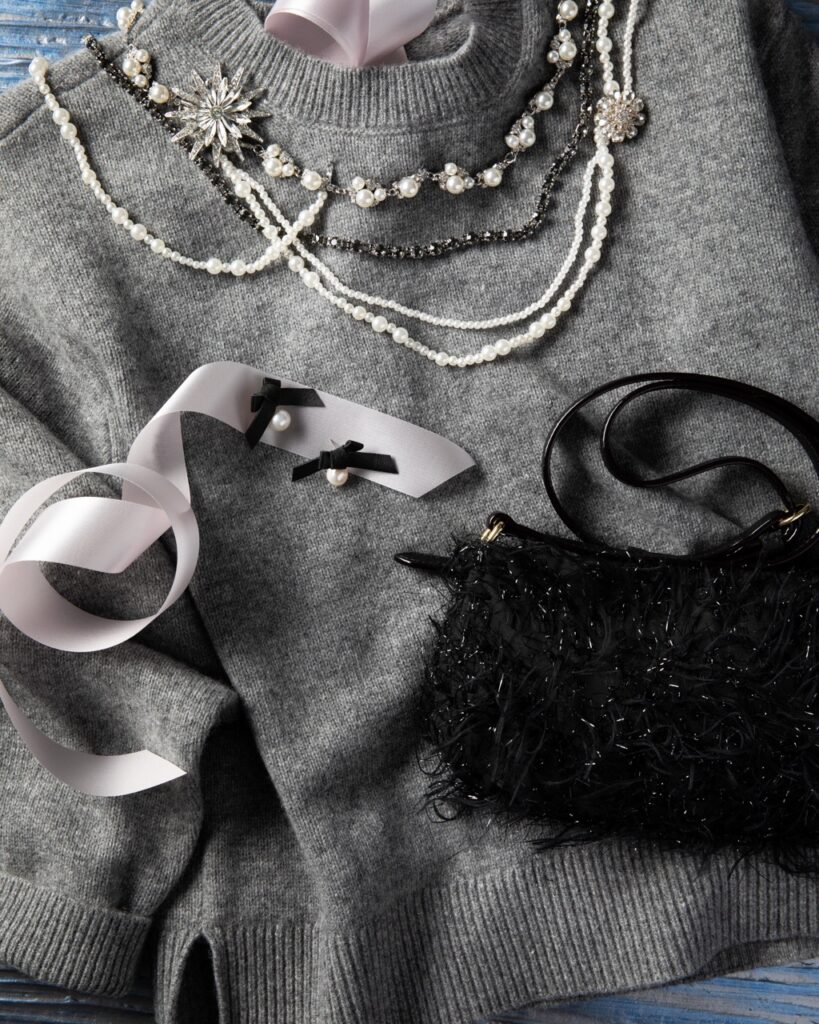 A grey sweater lays out with necklaces, earrings, a white ribbon, and a black purse laid on top of it.