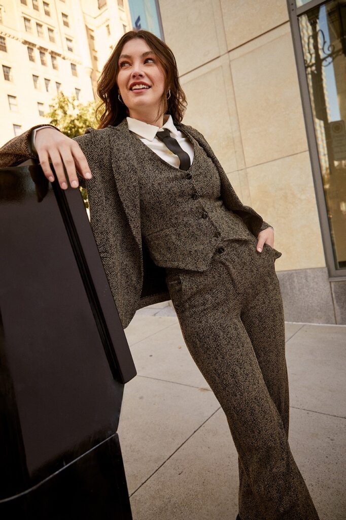 A woman leans against a stand with a full piece suit set in a dark brown twill style.