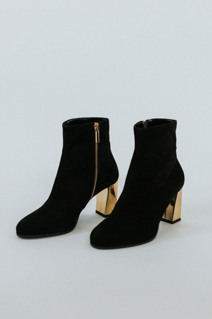 A pair of women's black ankle boots with gold heels sit on a white background.