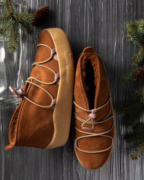 A pair of women's brown moccasins sit on a grey background with pine branches on the sides.