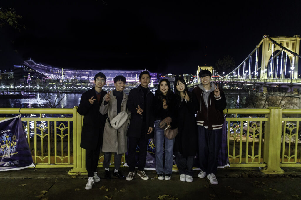 Six young adults standing side by side in front of the river with a view of the city night sky behind them. 