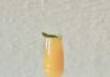 Pantone Color of the Year Peach Basil Bellini Cocktail & Mocktail A champagne flute with a peach cocktail garnished with a green basil leaf on a white surface with a champagne cork, basil leaves, and peach and orange flower petals for styling.
