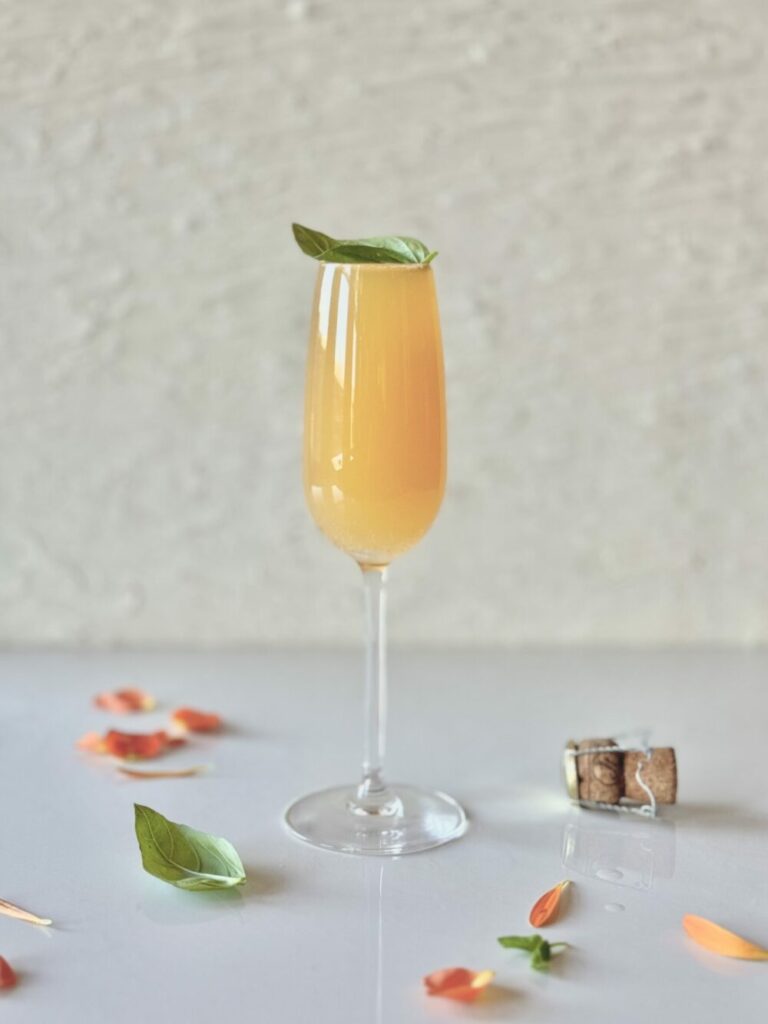 A champagne flute with a peach cocktail garnished with a green basil leaf on a white surface with a champagne cork, basil leaves, and peach and orange flower petals for styling.