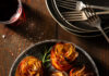 6 Holiday Sides by Chef Jackie Page A round crackled looking plate with 6 bacon potato roses garnished with fresh rosemary, a glass or red wine and a stack of little plates and forks.