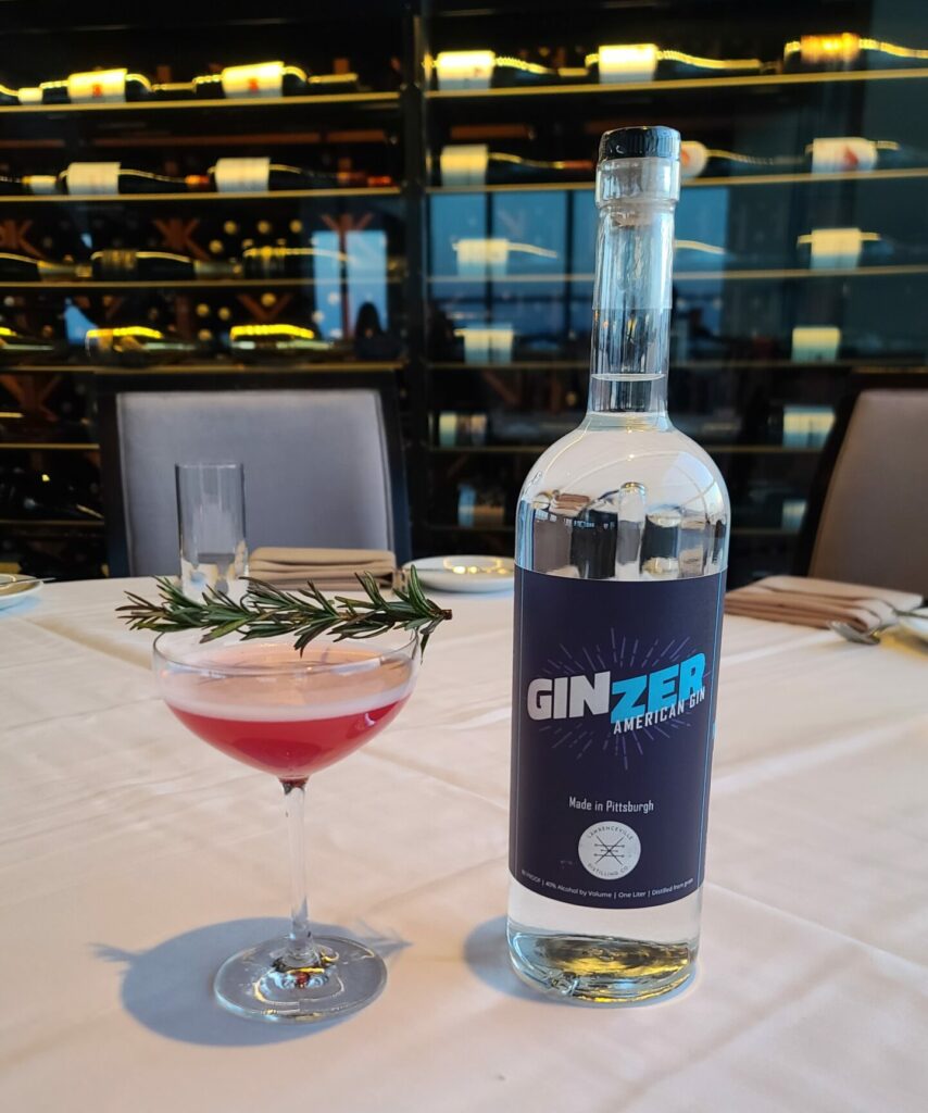 A photo of a martini glass filled with red liquid and topped with rosemary beside a bottole of GinZer gin on a table at Monterey Bay in Pittsburgh.