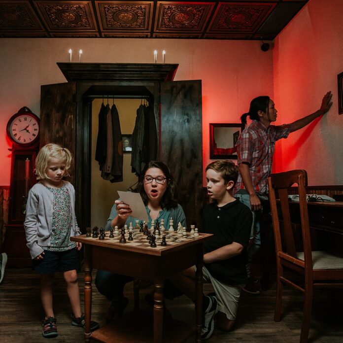 A family plays an escape game indoor activity in Pittsburgh as a mom, daughter, and son stand around a chess board and another person checks the wall behind them.