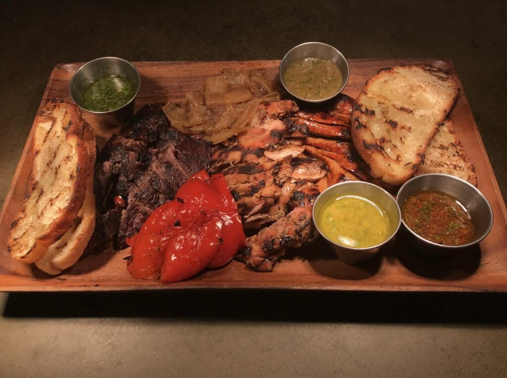 A wooden platter of various grilled meats, peppers, and sauces sits on a table at Gaucho in Pittsburgh.